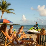 Pemandangan liburan di pantai oleh https://miami4d.com/ dengan teman-teman menikmati olahraga air saat senja.